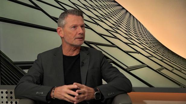 Mann mit kurzem grauem Haar trägt schwarzen Anzug und schwarzes T-Shirt, sitzt vor abstrakter Wand mit Gittermuster.