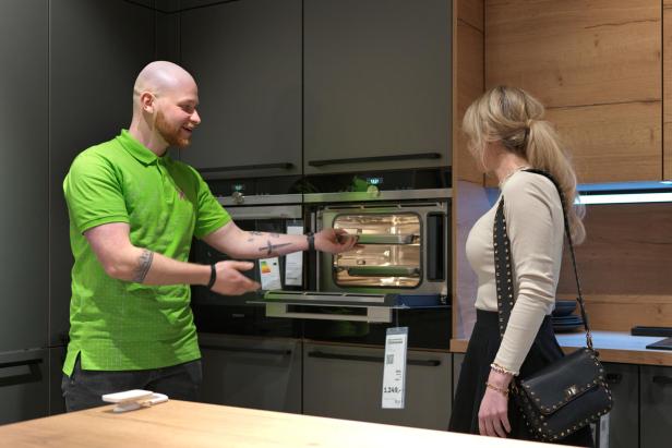 Mann in grünem Poloshirt zeigt Frau mit schwarzer Tasche einen geöffneten Backofen in moderner Kücheneinrichtung.