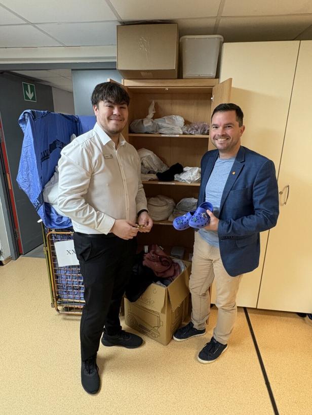 Zwei Männer stehen vor einem offenen Schrank mit Kleidung und einer Kiste, einer hält ein Paar blaue Gummischuhe in der Hand.