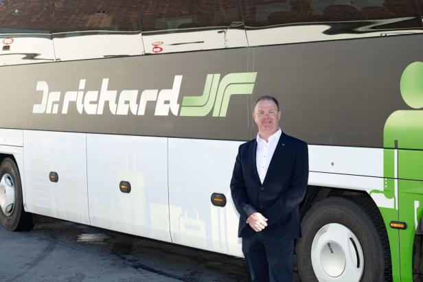 Ein Mann im Anzug steht lächelnd vor einem Reisebus mit der Aufschrift „Dr. Richard“.