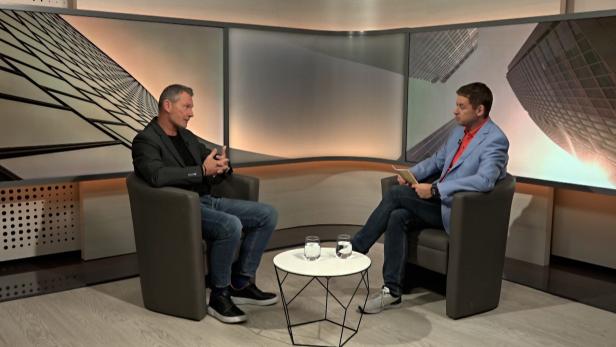 Zwei Männer sitzen sich in einem Studio gegenüber, beide in Sesseln, zwischen ihnen ein runder Tisch mit zwei Wassergläsern.