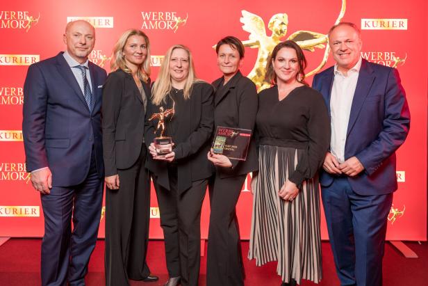 Sechs Personen in festlicher Kleidung posieren vor rotem Hintergrund mit Trophäe und Auszeichnung.