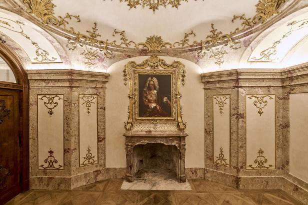 Prunkvoll verzierter Kamin mit goldgerahmtem Porträt eines Mannes in barockem Raum mit Stuck und Holzfußboden.