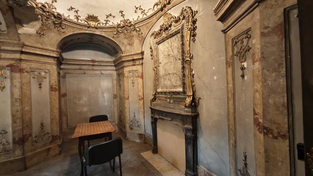 Prunkvoll verzierter Raum mit Kamin, goldenen Stuckelementen, einem kleinen Holztisch und zwei schwarzen Stühlen.