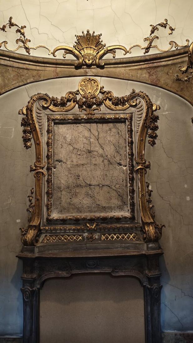 Prunkvoll verzierter Kamin mit goldenen Ornamenten und leerem, marmoriertem Rahmen an der Wand.