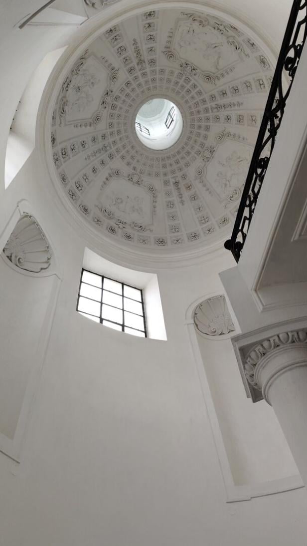 Weiße, reich verzierte Kuppeldecke mit Oberlicht und einem Fenster, daneben ein schmiedeeisernes Geländer.