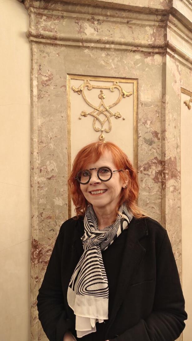 Eine lächelnde Frau mit roten Haaren, Brille und gemustertem Schal steht vor einer verzierten Marmorwand.