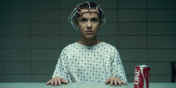 Ein Kind (Millie Bobbie Brown als Elf) mit kurzgeschnittenen Haaren trägt ein Krankenhaushemd und eine Elektrodenkappe, vor ihm steht eine Dose Cola.