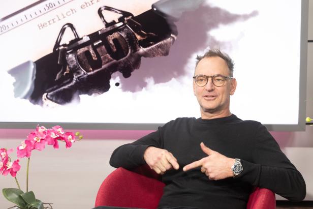 Ein Mann mit Brille sitzt auf einem roten Stuhl, im Hintergrund eine große Schreibmaschinenabbildung und eine pinke Orchidee.
