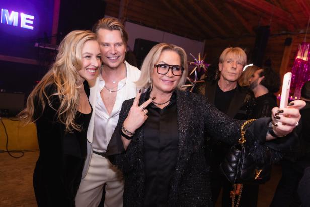 Drei fröhliche Menschen posieren für ein Selfie auf einer Party, während weitere Gäste im Hintergrund stehen.