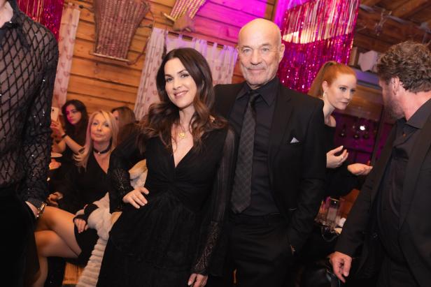 Mehrere elegant gekleidete Menschen feiern gemeinsam in einer gemütlich dekorierten Holzhütte mit pinker Beleuchtung.