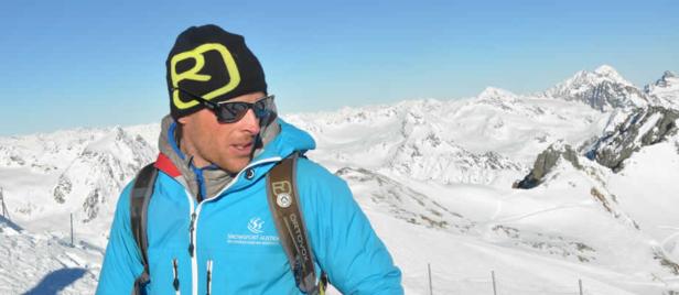 Person in blauer Jacke und schwarzer Mütze mit gelbem Muster vor schneebedeckten Bergen und blauem Himmel.