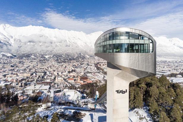 Rundes, modernes Aussichtsturmgebäude mit Glasfassade auf hohem, schmalem Sockel vor verschneiter Berglandschaft und Stadt im Tal.