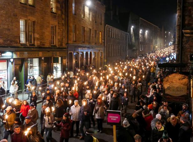 Eine Menschenmenge zieht nachts mit Fackeln durch eine belebte Straße in Edinburgh.