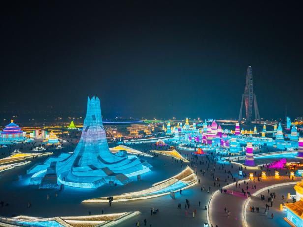 Bunte, beleuchtete Eisskulpturen und Gebäude in einer winterlichen Parklandschaft mit vielen Besuchern bei Nacht.