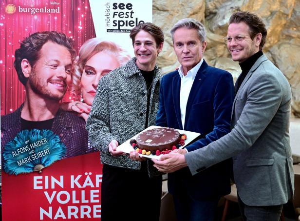 Drei Männer halten gemeinsam eine Schokoladentorte mit Früchten vor einem Plakat mit zwei Porträts und rotem Hintergrund.