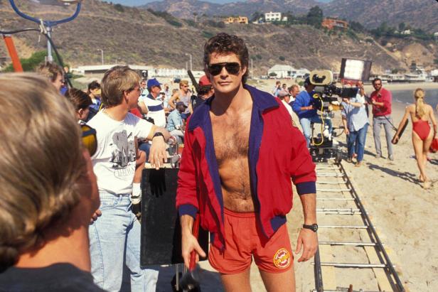 Drearbeiten am Strand: Hasselhoff steht mit Sonnenbrille lässig da, rund um ihn steht die Filmcrew.