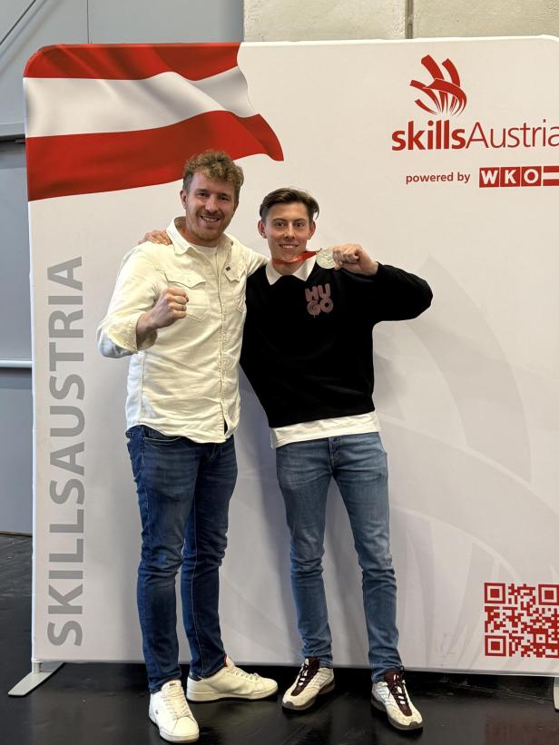 Zwei junge Männer jubeln vor einem SkillsAustria-Banner, einer hält eine Silbermedaille.