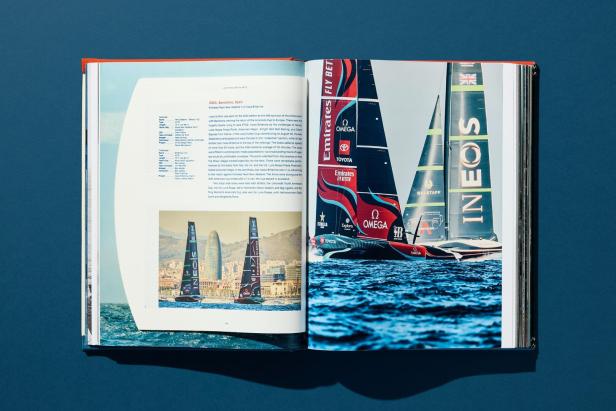 Zwei Segelboote mit auffälliger Werbung segeln auf dem Meer vor einer Stadt mit markantem Hochhaus im Hintergrund.