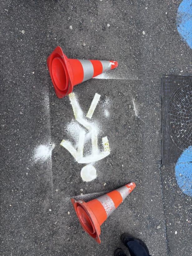 Zwei orange Leitkegel liegen auf einer Straße neben einer weißen Strichfigur auf dem Asphalt.