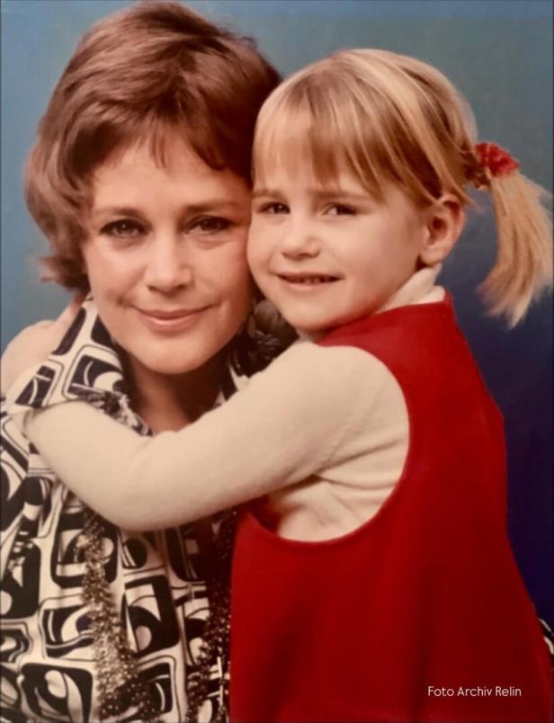 Eine Frau mit kurzen braunen Haaren wird von einem kleinen Mädchen mit blonden Zöpfen umarmt.