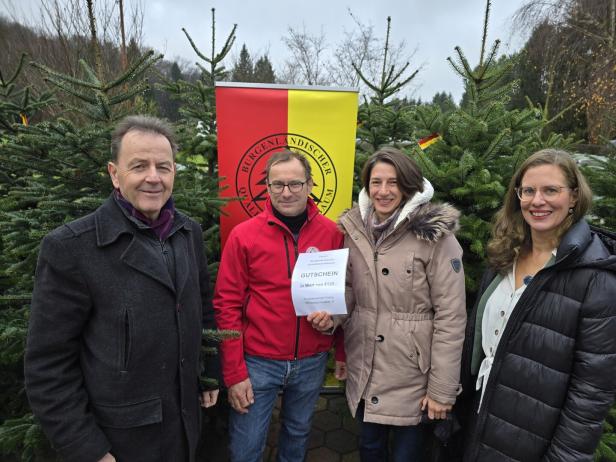 Vier Personen stehen lächelnd vor Tannenbäumen, eine Person hält einen Gutschein, im Hintergrund ist ein gelb-rotes Banner.