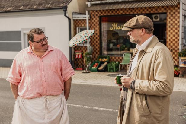 Zwei Männer unterhalten sich auf einer Straße vor einem kleinen Laden mit Gemüseauslage und Sonnenschirm.