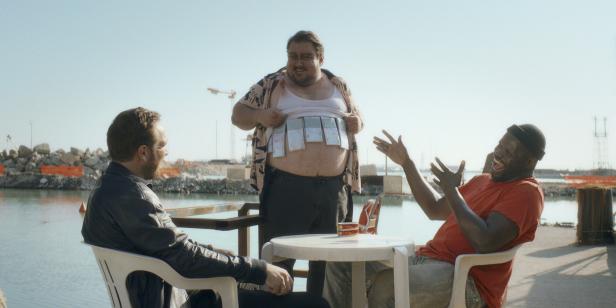 Drei Männer sitzen am Wasser, einer steht und zeigt lachend mehrere Geldscheine, die an seinem Bauch befestigt sind.