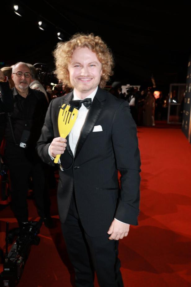 Paulus Bohl mit lockigem Haar und Smoking hält einen gelben Gegenstand auf einem roten Teppich.