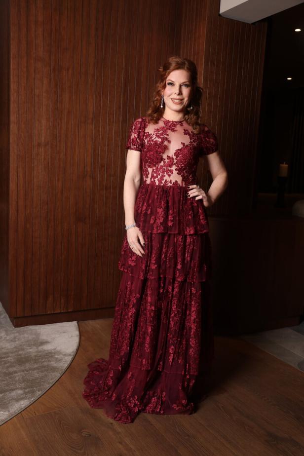 Frau mit langen, gelockten Haaren trägt ein bodenlanges, bordeauxrotes Spitzenkleid mit floralen Mustern.