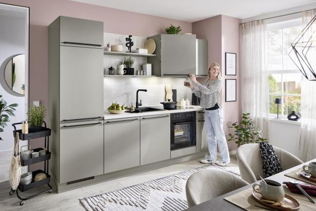 Eine Frau steht in einer modernen, hellen Küche und nimmt ein Glas aus einem Oberschrank, während Sonnenlicht durch das Fenster fällt.