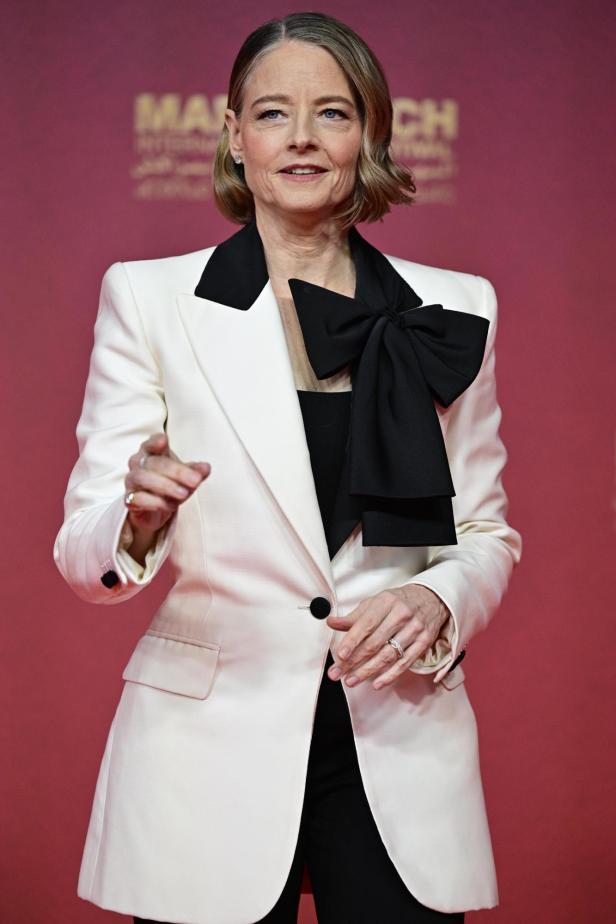 Jodie Foster trägt einen weißen Blazer mit schwarzem Revers und einer großen schwarzen Schleife vor einem roten Hintergrund.