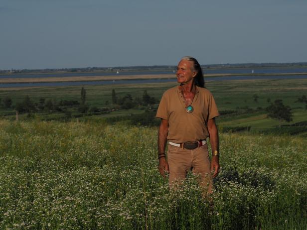 Ein älterer Mann steht in einem blühenden Feld und blickt lächelnd in die Ferne, im Hintergrund Flusslandschaft.