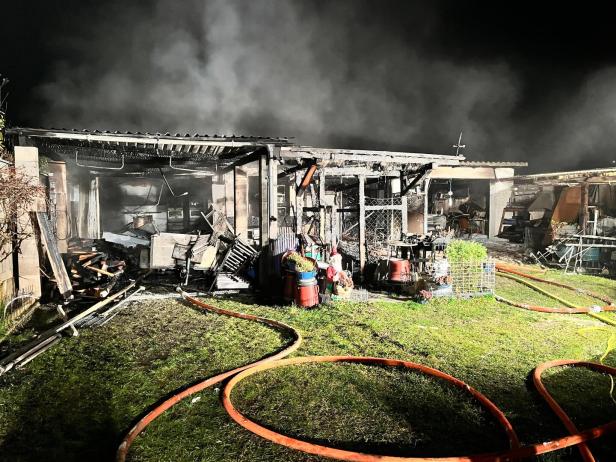 Ein ausgebranntes Gartenhaus mit Rauchentwicklung, Trümmern und Feuerwehrschläuchen auf dem Rasen.
