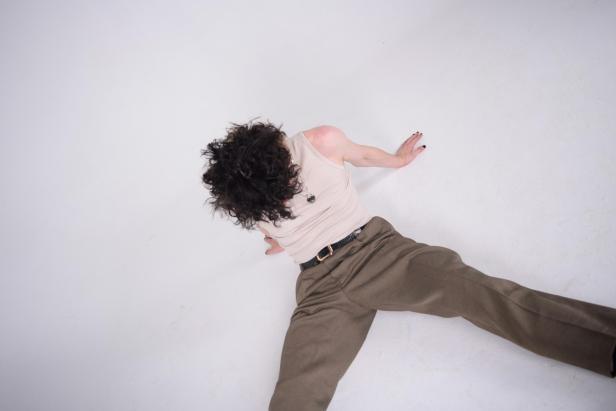 Person mit lockigen dunklen Haaren liegt nach einem Sturz auf hellem Boden, trägt ein helles Top und braune Hose.