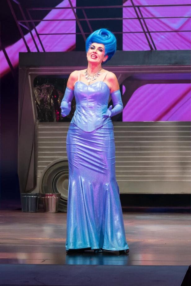 Eine Person mit blauer Hochsteckfrisur trägt ein glänzendes, bodenlanges Kleid und passende Handschuhe auf einer Bühne.