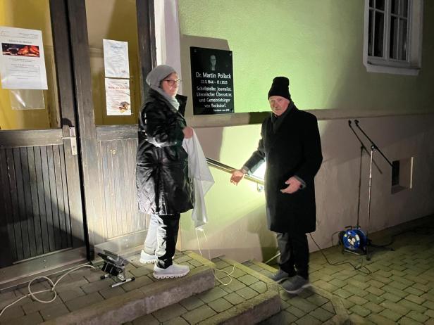 Zwei Personen enthüllen abends eine Gedenktafel an einer Hauswand neben einem Eingang.