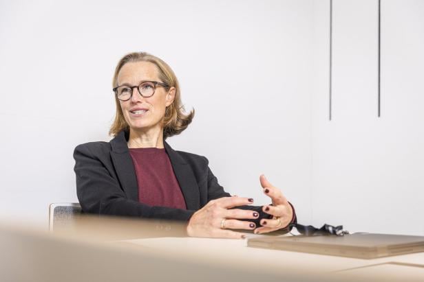 Frau mit Brille und dunklem Blazer sitzt an einem Tisch und gestikuliert beim Sprechen.