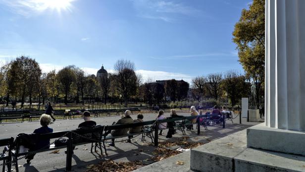 An einem sonnigen Tag sitzen Menschen auf Bänken in einem Park.