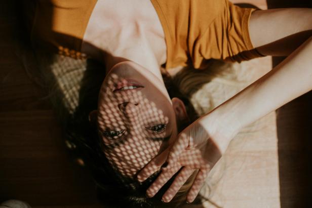 Frau liegt auf dem Rücken, trägt ein senffarbenes Top, Schattenmuster aus Gitter auf Gesicht und Arm.