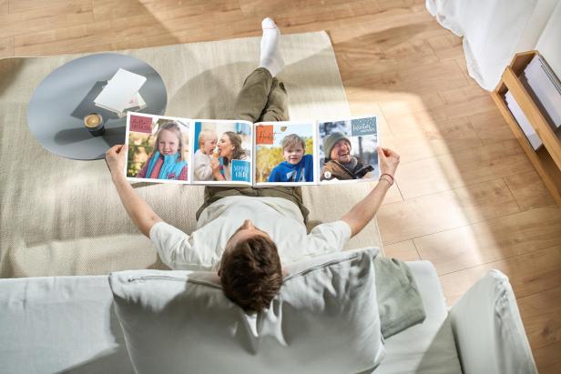 Person betrachtet entspannt ein großformatiges Fotobuch mit Familienfotos auf dem Sofa.