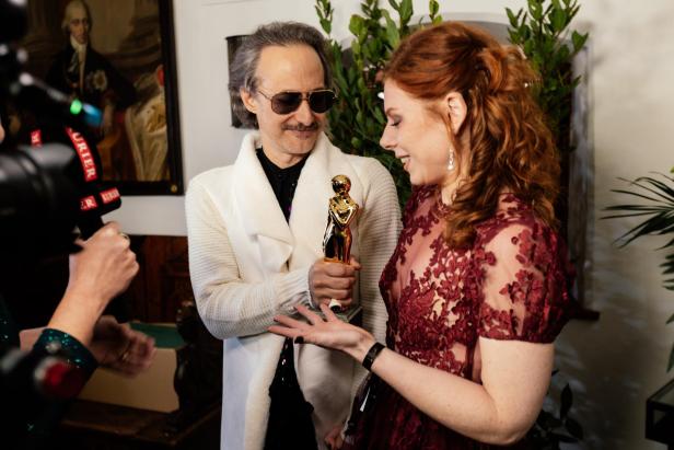 Ein Mann überreicht einer lächelnden Frau in elegantem Kleid eine goldene Trophäe, während eine Kamera sie filmt.