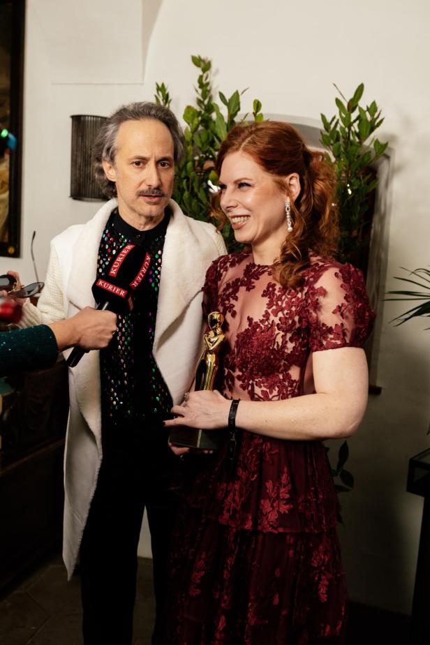 Eine Frau im roten Spitzenkleid hält eine goldene Trophäe, neben ihr steht ein Mann, während ein Mikrofon auf sie gerichtet ist.