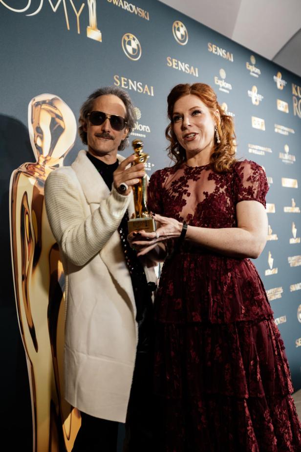 Ein Mann und eine Frau halten gemeinsam eine goldene Trophäe vor einer Sponsorenwand.