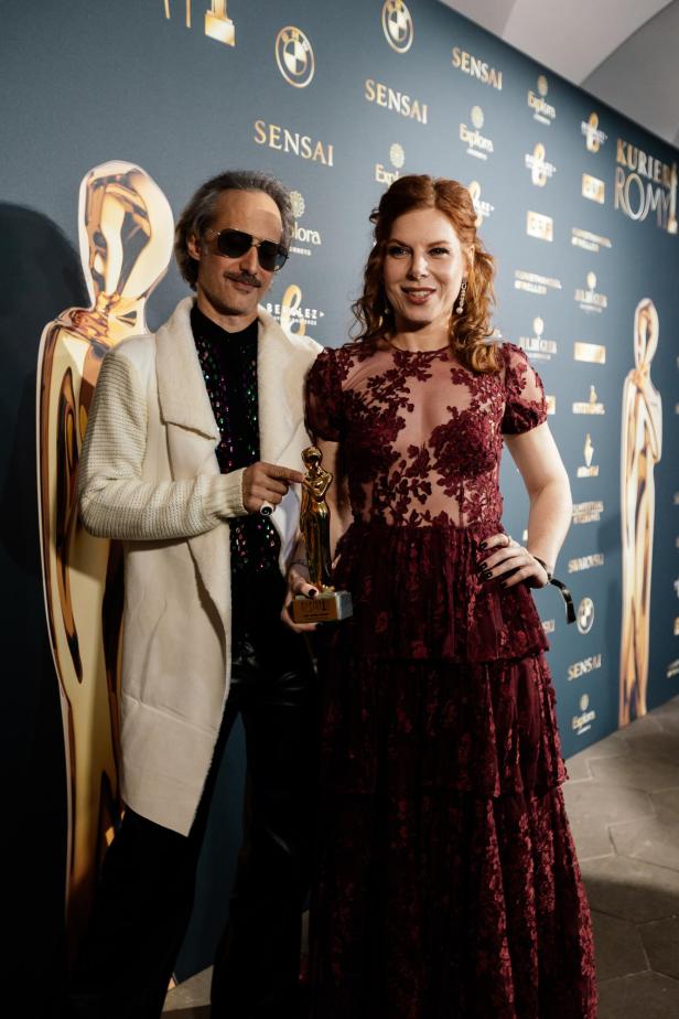 Ein Mann und eine Frau posieren elegant vor einer Wand mit Logos, die Frau hält eine goldene Trophäe in der Hand.