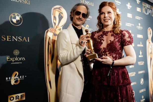 Zwei elegant gekleidete Personen halten gemeinsam eine goldene Trophäe und lächeln vor einer Sponsorenwand.