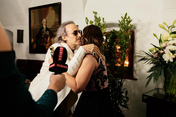 Ein Mann umarmt eine Frau, während eine Person mit einem KURIER-Mikrofon danebensteht und Blumen im Hintergrund stehen.