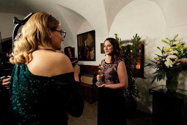 Eine Frau hält eine goldene Trophäe und wird von einer Reporterin mit Mikrofon interviewt, im Hintergrund Blumen und Gemälde.