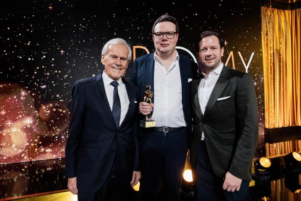 Drei Männer in Anzügen stehen vor einem funkelnden Hintergrund, einer hält eine goldene Trophäe in der Hand.