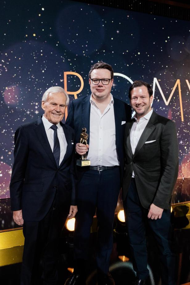 Drei Männer in Anzügen stehen lächelnd zusammen, einer hält eine goldene Trophäe vor funkelndem Hintergrund.